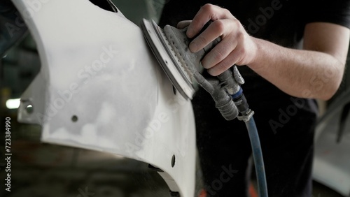 A worker professionally cleans the surface of a car bumper with a grinding machine in a car service garage before painting, close-up. Sanding a car bumper before painting.