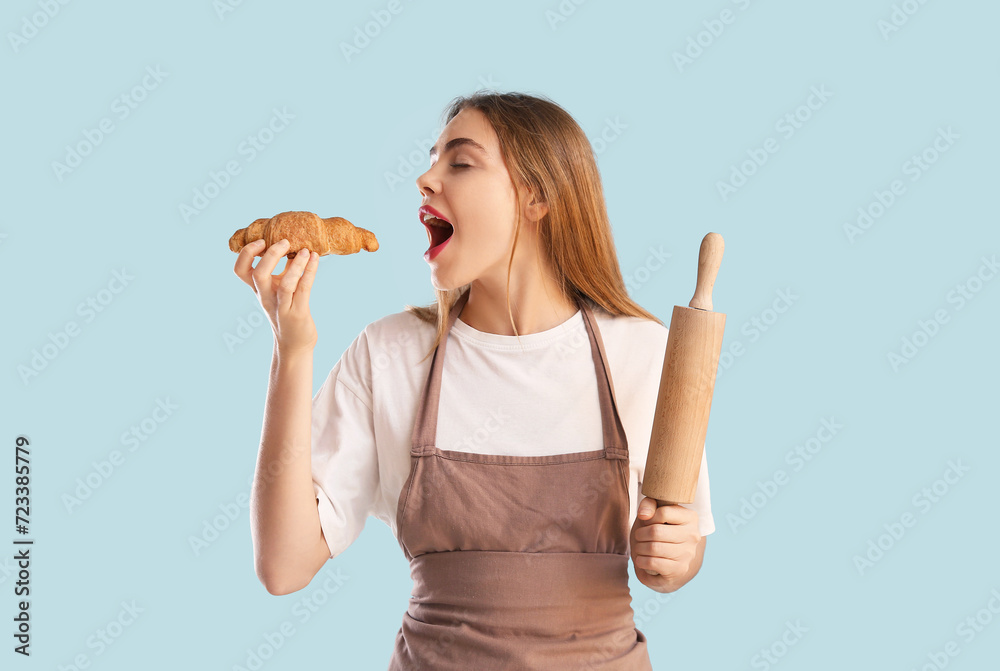 Pretty young woman in apron with rolling pin eating croissant on blue background Pretty young woman in apron with rolling pin eating croissant on blue background