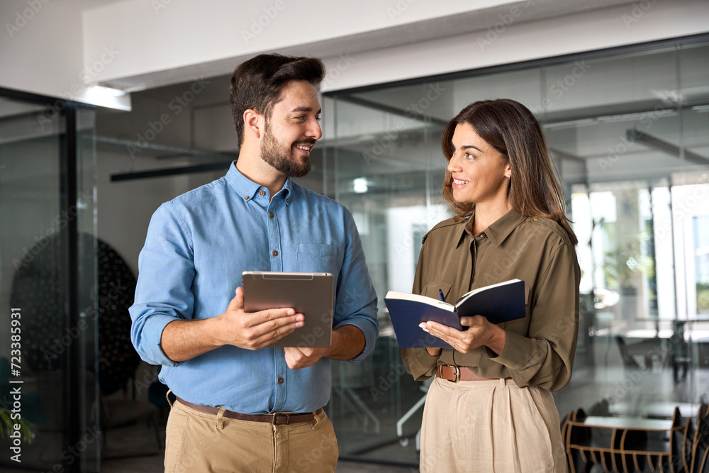 Happy busy latin partners using digital tablet talking standing in office. Professional business people having conversation sharing ideas. Female executive manager working together with male employee. Happy busy latin partners using digital tablet talking standing in office. Professional business people having conversation sharing ideas. Female executive manager working together with male employee.