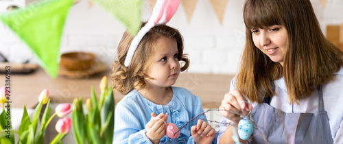 Mother and daughter painting eggs, creating home Easter decor together, happy, cheerful. Cute pretty smiling little girl, family time. Fresh spring bright colourful flowers in the kitchen. Banner