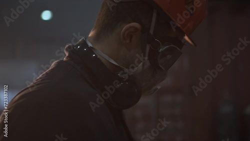 an unrecognizable worker in a hard hat and mask with reflections of light from sparks on his face