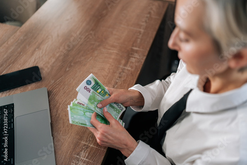Top view of happy rich business woman counting heap euro dollars sitting office desk in elegant wear. Close-up of female hands holding hundred banknotes European currency on pile. Concept of wealth