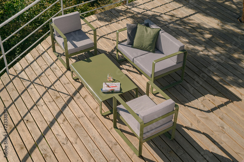 Summer terrace with a wooden floor, in the garden with a sofa, two armchairs and a table, a magazine and a glass on the table