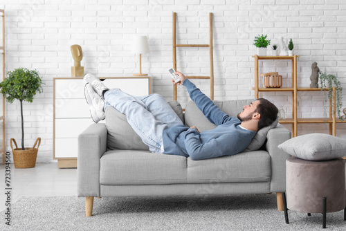 Handsome young man with air conditioner remote control lying on sofa in living room photo