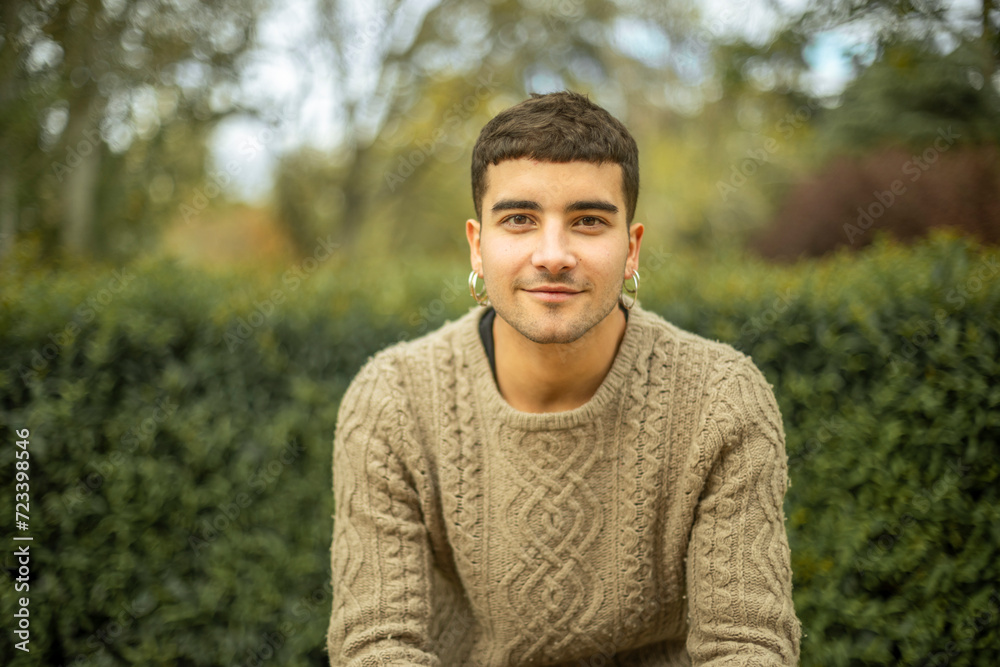 Outdoor portrait of young caucasian man with earrings smiling with blurred plant background looking at camera