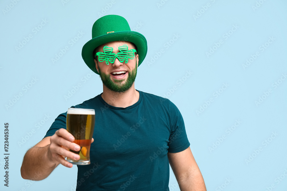 Young man in leprechaun hat and decorative glasses in shape of clover with green beard holding glass of beer on blue background. St. Patrick's Day celebration Young man in leprechaun hat and decorative glasses in shape of clover with green beard holding glass of beer on blue background. St. Patrick's Day celebration