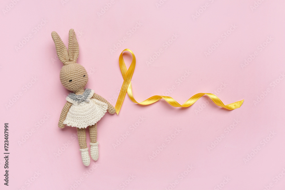 Golden ribbon with toy bunny on pink background. Childhood cancer awareness concept - obrazy, fototapety, plakaty 