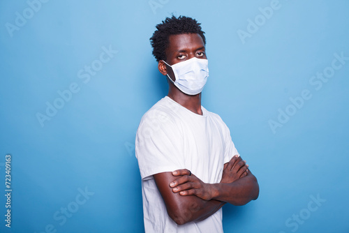 Portrait of black person with face mask standing with arms crossed while looking at camera. African American man with healthcare protection against coronavirus pandemic. Young guy in studio. photo
