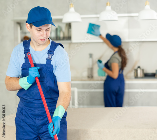 Guy employee of cleaning company in blue jumpsuit cleans and washes floor in kitchen and together with girl assistant cleans surfaces and furniture