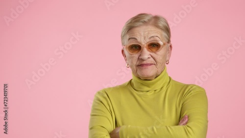Well-groomed elederly lady with blonde short hair in stylish spectacles, yellow turtleneck standing over pink background, looking with disrespect, contempt. High quality 4k footage