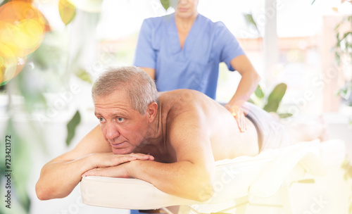 Relaxed old man undergoing back massage therapy conducted by skilled masseuse in light room