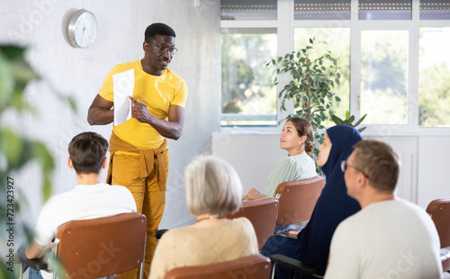Portrait of confident african american tutor giving lecture to multiethnic group of adult students  standing in front of audience with papers in hands..