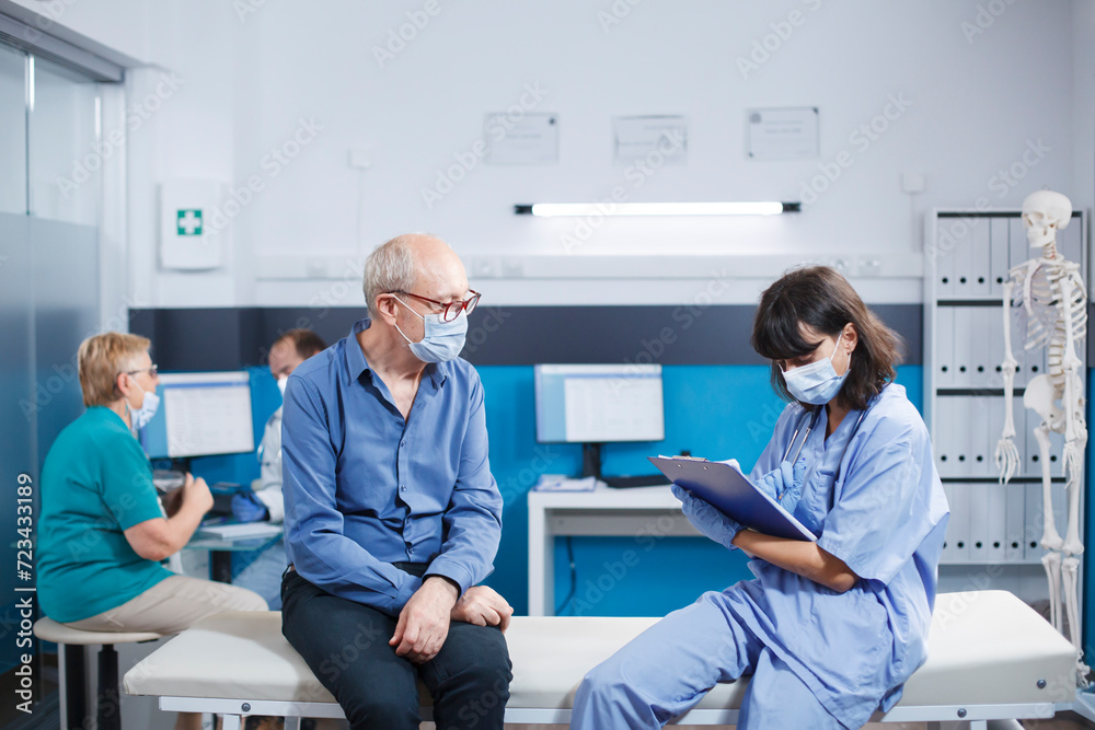 Nurse practitioner taking notes at consultation with senior man in hospital office. Elderly male patient attending healthcare examination with medical assistant during covid 19 pandemic. Nurse practitioner taking notes at consultation with senior man in hospital office. Elderly male patient attending healthcare examination with medical assistant during covid 19 pandemic.