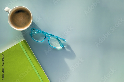A morning coffee mug, a pair of blue reading glasses and a green notebook on a solid blue background