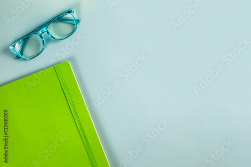 A pair of blue reading glasses and a green notebook on a solid blue background with empty copy space