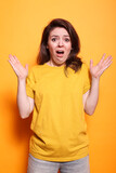 Frightened brunette lady showing face expression of despair, standing over orange background. Shocked woman feeling scared and afraid with hands up while looking at camera.