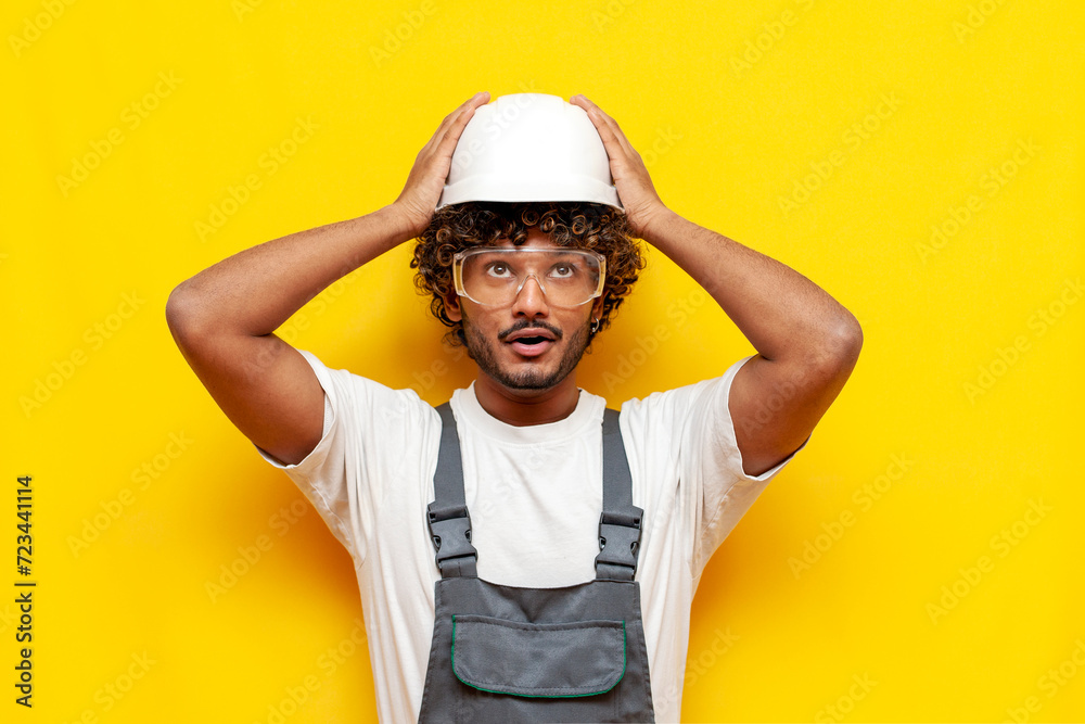 A young Indian builder in safety glasses and overalls puts a hard hat on his head on a yellow isolated background, a Hindu foreman in uniform holds a white hard hat and looks up in surprise