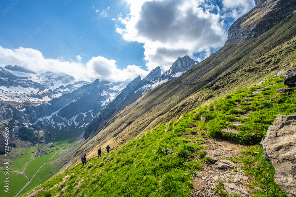 Fototapeta premium Tourists walk along a hiking trail through a green meadow with an alpine valley and a steep, rocky mountain formation in the background. Seealpsee, Appenzell, Switzerland, Europe.
