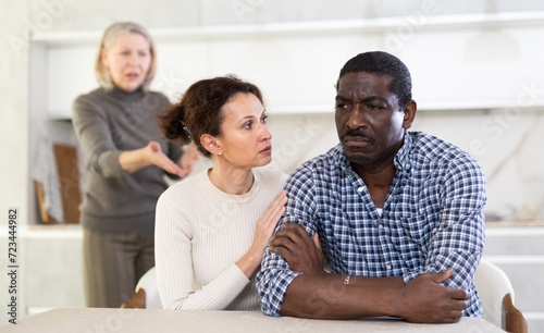 Middle-aged woman trying to calm angry man sitting at the kitchen table while mature woman quarreling to him standing behind