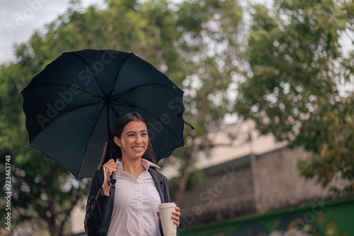 A beautiful smiling Latin woman walks down the street on a rainy day with an umbrella and a coffee in her hand.
