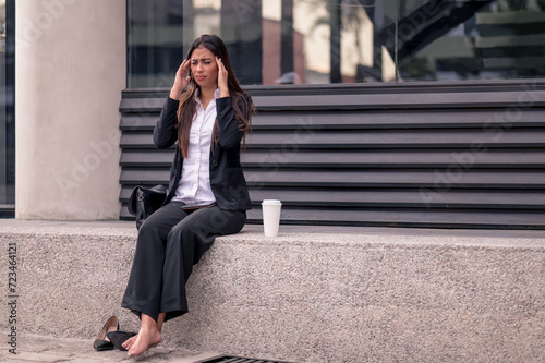 Business woman sitting on the street with headache. High shoes and a formal suit massages her head.