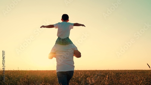 Little kid superhero imagines soaring through sky at sunset. Loving father runs with playful son on shoulders through wheat field. Happy child daydreams about pilot gliding in golden glow of sunset