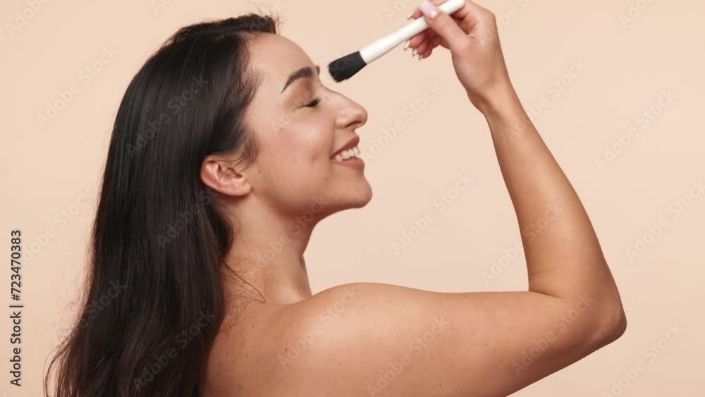 Beautiful brunette young female smiling as she uses a makeup brush on her nose and face against a beige background, depicting beauty, skincare, and happiness. Beautiful brunette young female smiling as she uses a makeup brush on her nose and face against a beige background, depicting beauty, skincare, and happiness.