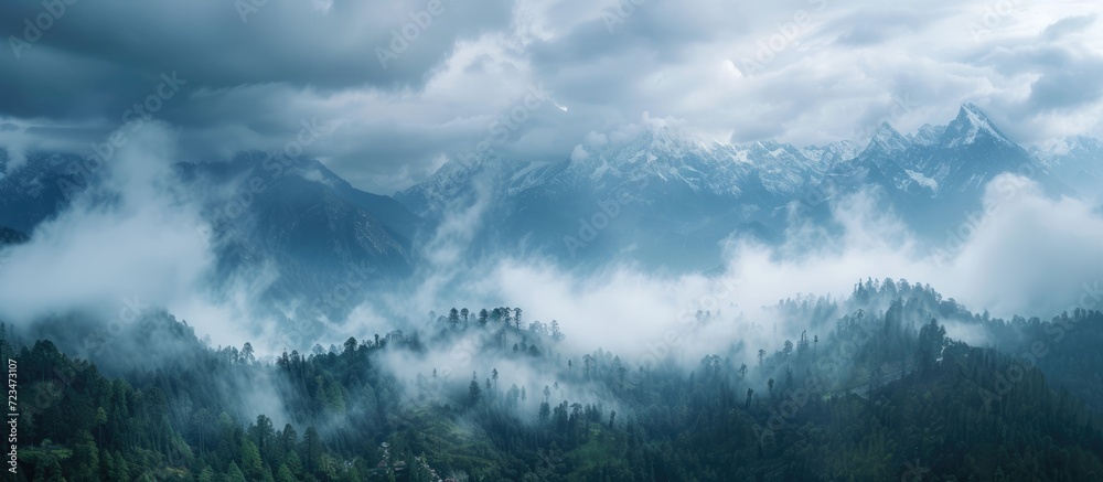 Fototapeta premium From above, the contrasting beauty of nature is seen: clouds veiling majestic peaks, the sky peeking through, and a mix of forest and clear-cuts in the foreground while gray clouds approach.