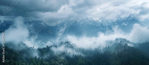 From above, the contrasting beauty of nature is seen: clouds veiling majestic peaks, the sky peeking through, and a mix of forest and clear-cuts in the foreground while gray clouds approach. © Vusal