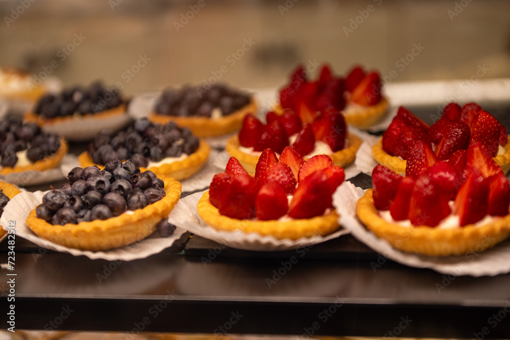 A tray of fruit filled custard tarts. The toppings are blueberries, and strawberries on an almond crust. The glass display case has both strawberry and blueberry mini tarts on white paper doilies. A tray of fruit filled custard tarts. The toppings are blueberries, and strawberries on an almond crust. The glass display case has both strawberry and blueberry mini tarts on white paper doilies.