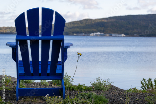 A royal blue wooden Adirondack chair on rich green grass overlooking the calm blue ocean under a cloudy blue sky.  There's a rocky shoreline in the distance with trees, shrubs, and red foliage.  photo