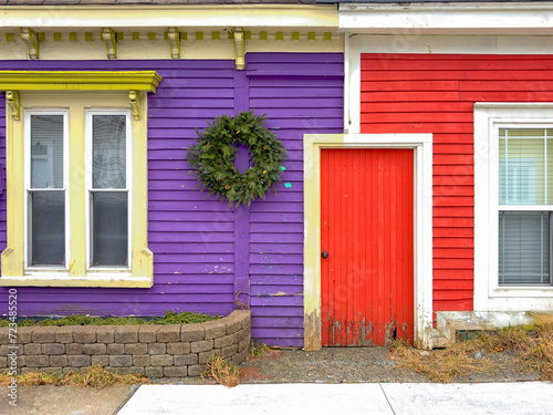 A vibrant red wooden house with a shared small red door to a bright purple building. There's a green wreath hanging from the exterior wall. The wood trim is cream color. There are double-hung windows.