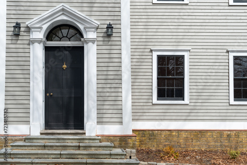 The exterior of a vintage building with beige-colored narrow clapboard Cape Cod siding. There's a black wooden door with a thick white decorative trim. A half-circle transom window hangs over the door