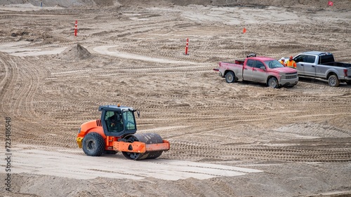 Vibratory soil roller compactor compacting a thickness of sand on a construction site, with two pick-ups and workers