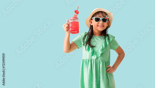 Happy Asian little girl posing with wear a hat and sunglasses holding glass of red sweet water, Holiday summer fashion green dress, isolated on pastel blue color background
