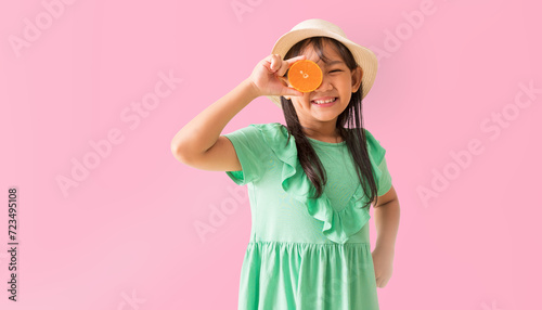 Happy Asian little girl posing with wear a hat with sunglasses holding orange slices on face, Holiday summer fashion green dress, isolated on pastel pink color background