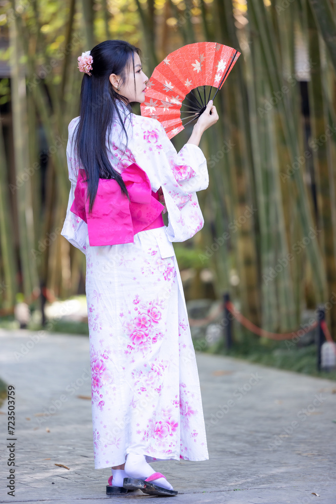 Asian woman wearing japanese traditional kimono standing on street. Beautiful Woman smiling and holding hand fan to cover face. Woman in Kimono dress enjoying and walking on bamboo forest. Asian woman wearing japanese traditional kimono standing on street. Beautiful Woman smiling and holding hand fan to cover face. Woman in Kimono dress enjoying and walking on bamboo forest.