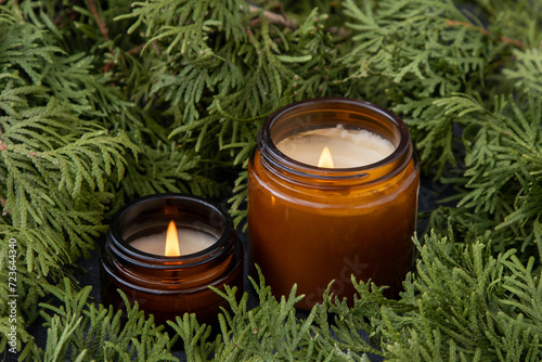 Soy candle in a glass jar. Green thuja branches background. Aromatherapy at home. Scented candles decor. photo