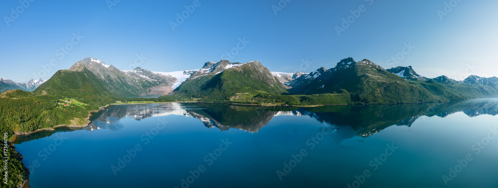 A panoramic view captures the serene beauty of Svartisen Glacier nestled in the Saltfjell mountains, with a pristine lake reflecting the rugged peaks under a clear blue sky