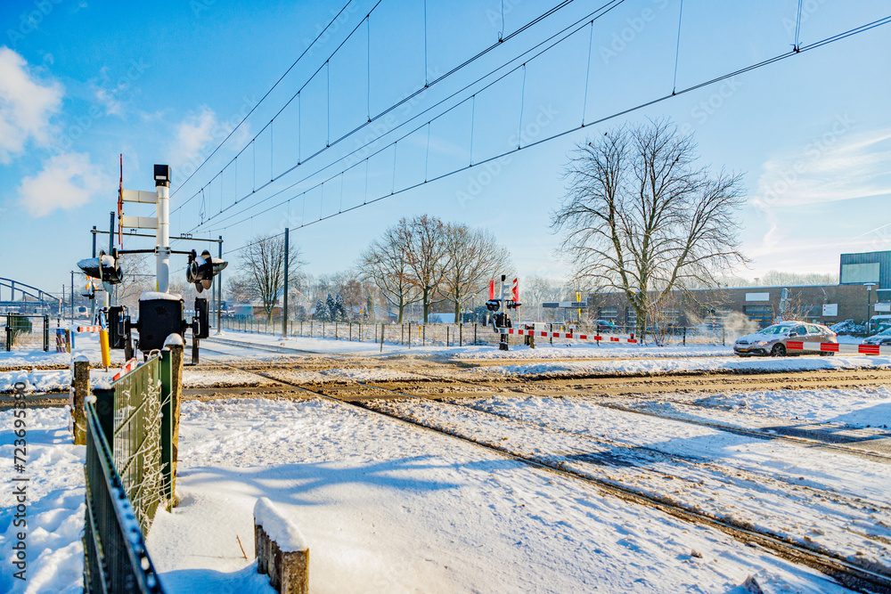 Snow covered railway crossing on road in winter landscape, closed downward barriers, stop sign and warning lights, stopped car waiting in background, sunny day in Beek, South Limburg, Netherlands