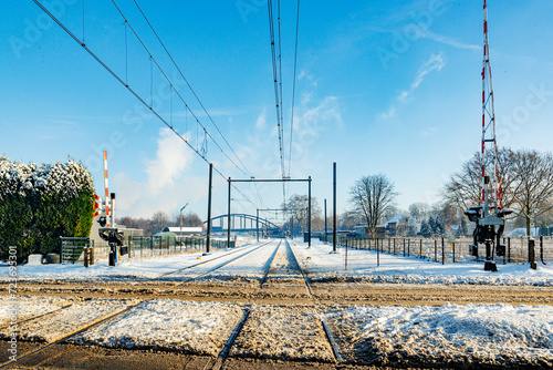 Snow covered train tracks at railway crossing, raised protective barriers, electric cables, bridge in foggy background, sunny day after heavy snowfall in Beek - Elsloo, South Limburg, Netherlands photo