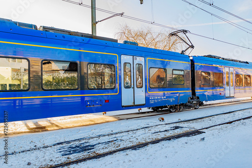 Blue carriage of an electric passenger train running on snow-covered tracks, sunny day after a heavy snowfall in Beek - Elsloo, South Limburg, Netherlands