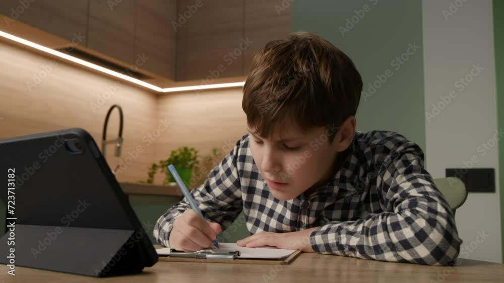 Diligent schoolboy writes with a pen, immersed in online homework, glancing at his tablet. A glimpse into modern study methods and tech-integrated learning Diligent schoolboy writes with a pen, immersed in online homework, glancing at his tablet. A glimpse into modern study methods and tech-integrated learning