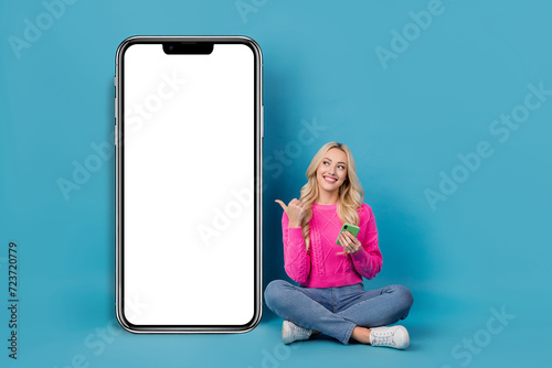 Full body photo of sitting girl crossed legs direct finger look mockup phone board smile menu beauty shop isolated on blue color background © deagreez