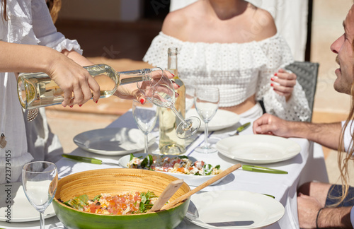 Close-up of a hand pouring white wine into a glass at a sunlit outdoor dining table, with a fresh salad in the foreground and friends gathered around, embodying the essence of a summer meal.