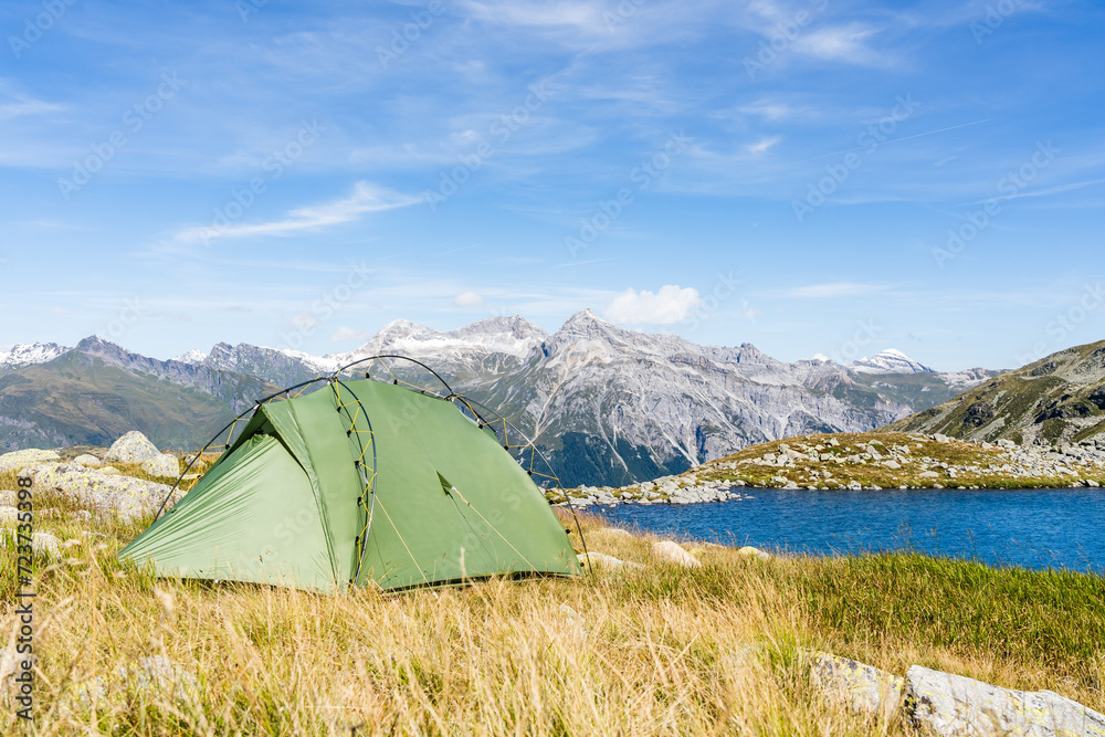 Tent in the grass next to a small alpine lake on a mountain pass in Switzerland on a sunny summer day