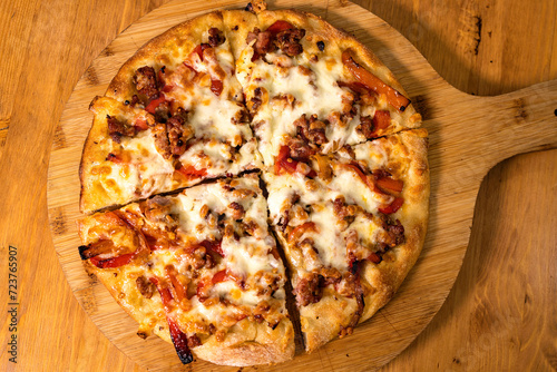 Pizza rotonda cotta nel ruoto condita con mozzarella, peperoni e salsiccia poggiata su un tagliere di legno di una rosticceria photo