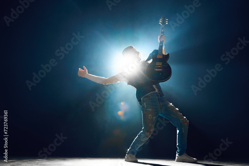 Rock performer in deep stance playing electric guitar on stage with backlit and smoke which creating his silhouette. Concept of Rock-n-roll, music and dance, festivals and concerts, culture.