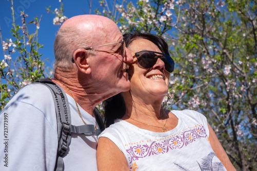 Portrait of happy smiling senior couple exchanging kisses and cuddling in natural park with flowering almond trees. Spring and almond blossoms are blooming.