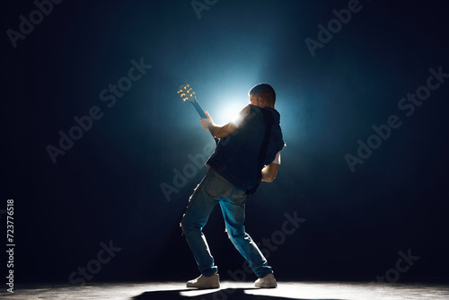 Performer with electric guitar leaning into musical moment against dark background with spotlights behind him. Concept of Rock-n-roll, music and dance, festivals and concerts, culture. photo